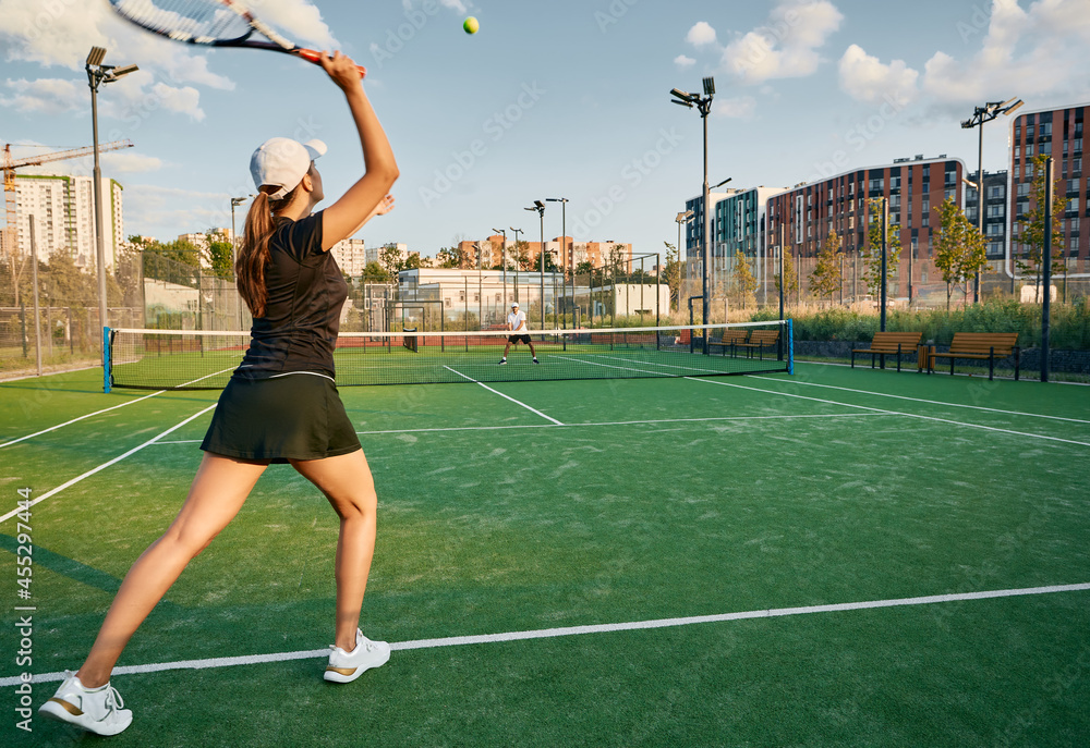 Tennis player serves ball while playing match with her male partner on a grass court in urban environment. Female tennis player with tennis racket and ball in action