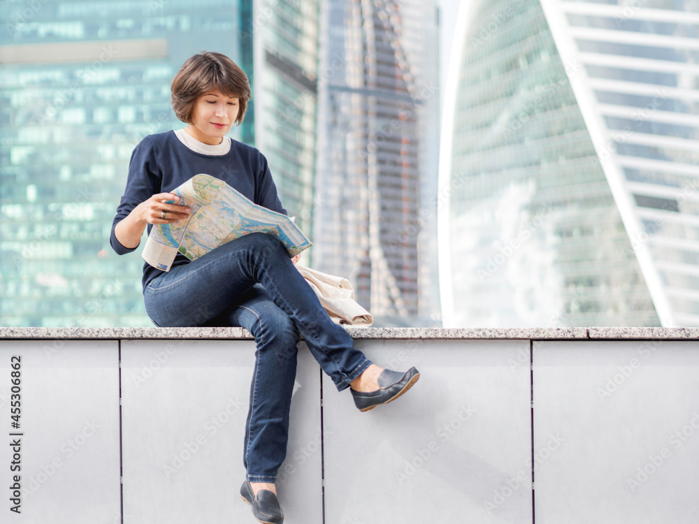 Smiling woman is reading paper map sitting on background of buildings with glass walls. Travel around city. Urban tourism. Modern architectural landmarks.