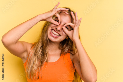 Young caucasian blonde woman isolated on yellow background showing okay sign over eyes