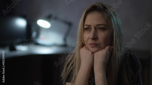 Stressed blonde woman with messy blonde hair suffers from depression and negative emotions in semi-dark room with glowing lamp extreme close view slow motion