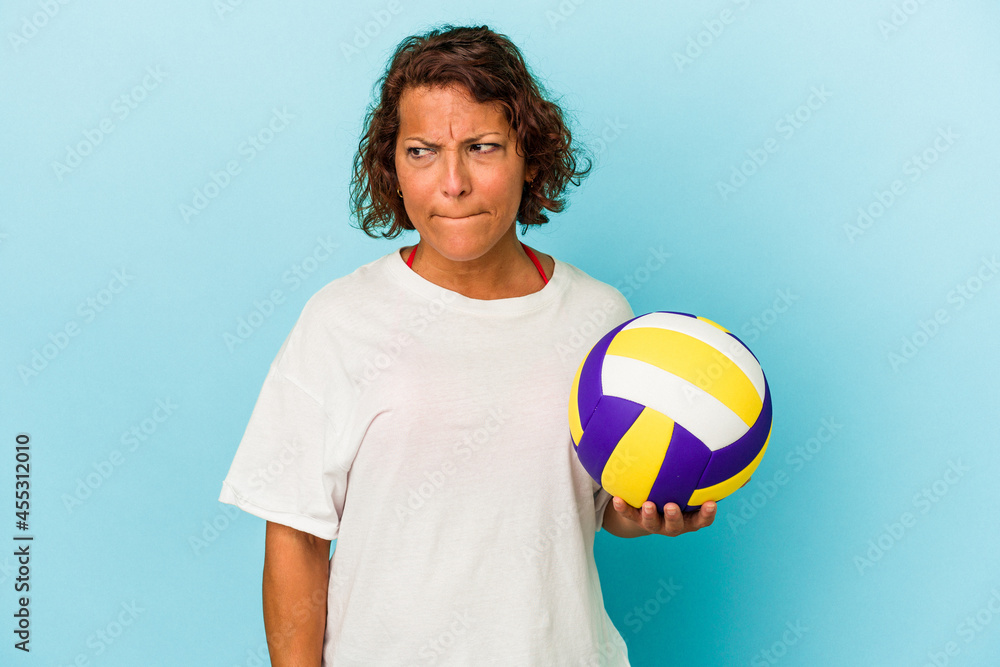 Middle age latin woman playing volleyball isolated on blue background confused, feels doubtful and unsure.