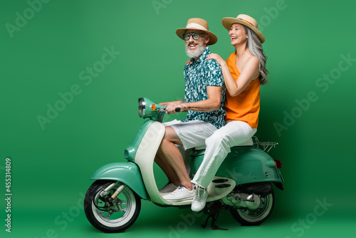 cheerful interracial middle aged couple in sun hats riding moped on green