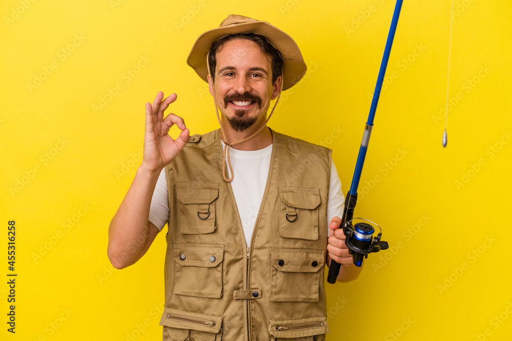 Young caucasian fisherman holding rod isolated on yellow background cheerful and confident showing ok gesture. Young caucasian fisherman holding rod isolated on yellow background cheerful and confident showing ok gesture.