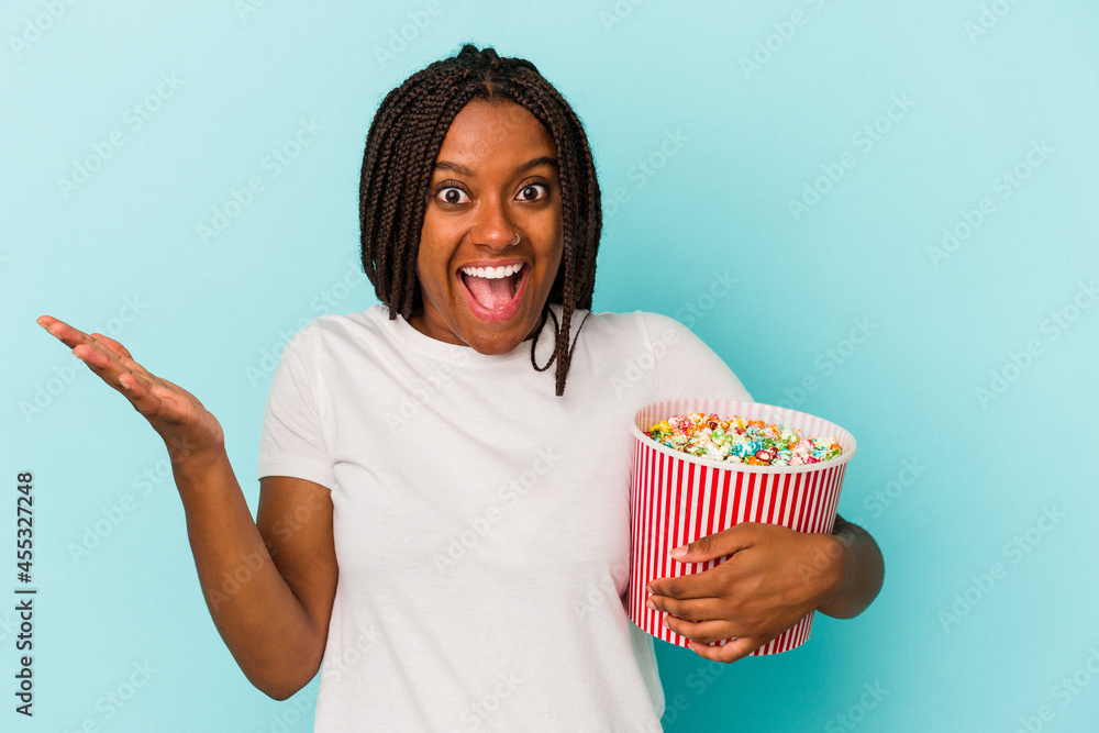 Young African American woman eating pop corns isolated on blue background  receiving a pleasant surprise, excited and raising hands.