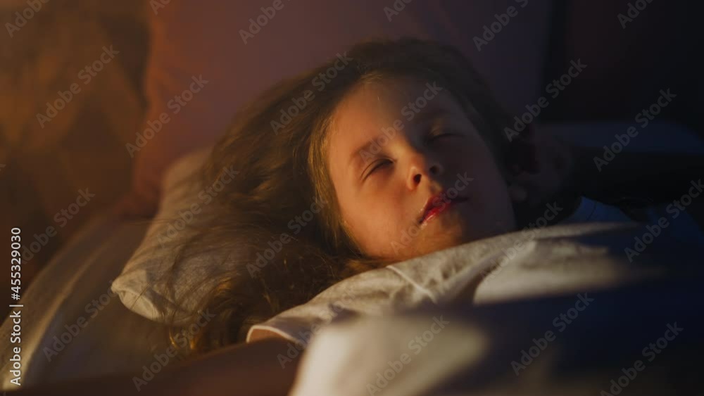 Sweet little girl sleeps with eyes blinking under closed eyelids covered with soft duvet on bed at gentle light of night lamp in dark room slow motion closeup Sweet little girl sleeps with eyes blinking under closed eyelids covered with soft duvet on bed at gentle light of night lamp in dark room slow motion closeup