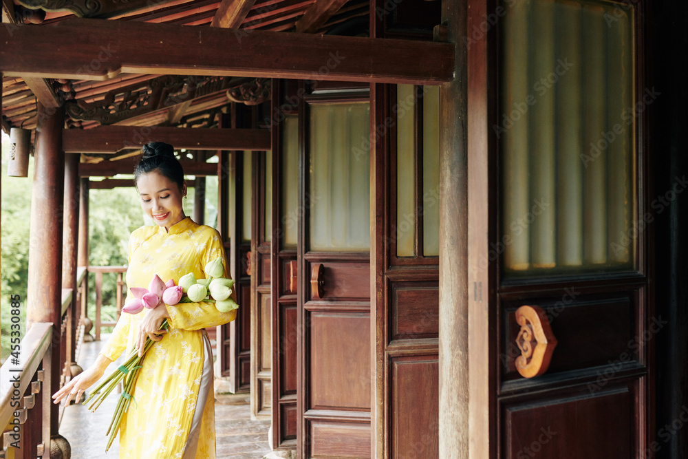 Beautiful smiling young woman in ao dai dress standing on porch with bunch of lotus flowers Beautiful smiling young woman in ao dai dress standing on porch with bunch of lotus flowers