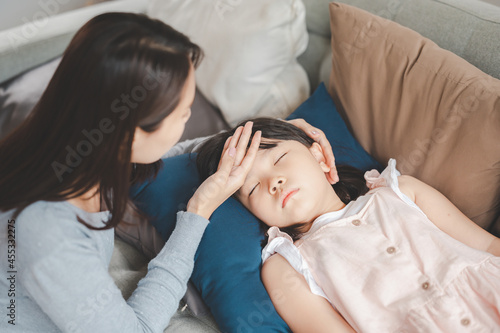 Sick Asian girl daughter sleeping on the couch with fever while mother checking temperature on her forehead
