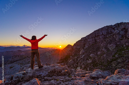 Happy hiker girl meets sunrise with raised arms high in the mountains.