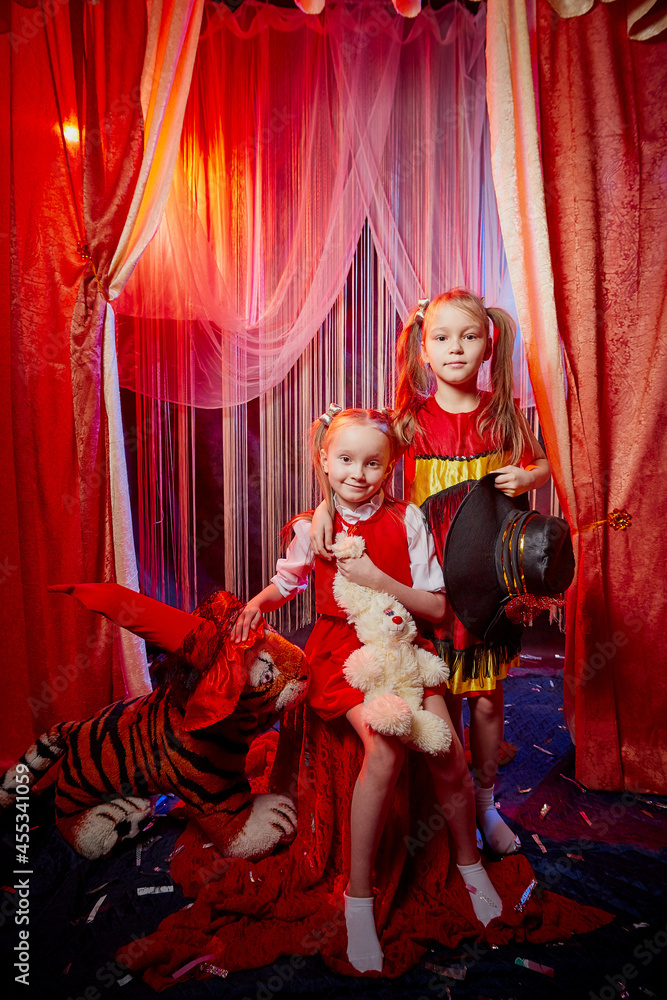 Small girls during a stylized theatrical circus photo shoot in a beautiful red location. Young models posing on stage with curtain. Twin sisters or female friends together