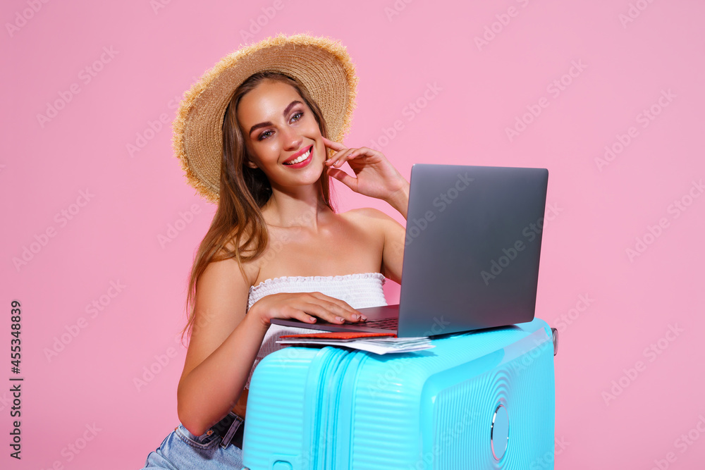 Smiling girl with laptop tickets money and passport are going to travel. Portrait. Sitting near suitcase in shorts white top and straw hat. Pink background. looks at the camera Smiling girl with laptop tickets money and passport are going to travel. Portrait. Sitting near suitcase in shorts white top and straw hat. Pink background. looks at the camera