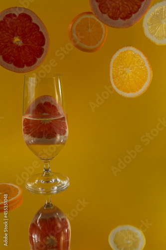 A closeup shot of a glass of water on a specular surface and orange, lemon and grapefruit slices against a yellow background