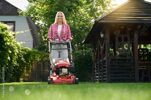 Obraz na plátně Beautiful young woman in casual clothes and protective gloves trimming grass with lawn mower during warm sunny day