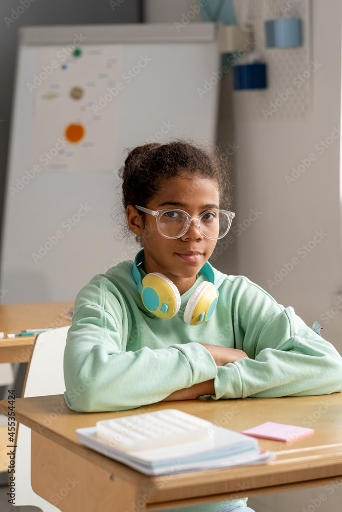 Cute serious schoolgirl in eyeglasses sitting by her desk and looking at camera at lesson Cute serious schoolgirl in eyeglasses sitting by her desk and looking at camera at lesson