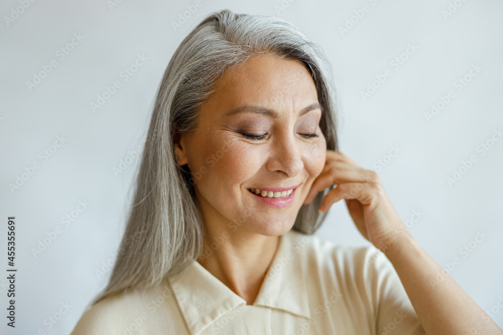 Attractive smiling grey haired lady in stylish shirt touches cheek by hand on light background in studio. Mature beauty lifestyle Attractive smiling grey haired lady in stylish shirt touches cheek by hand on light background in studio. Mature beauty lifestyle