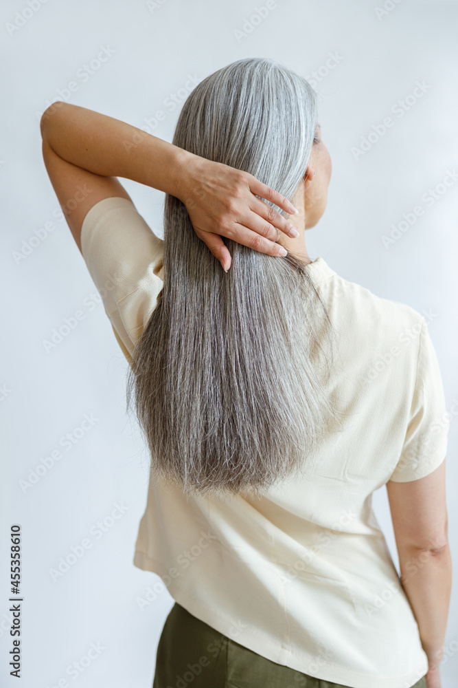 Fototapeta premium Middle aged woman in yellow blouse adjusts long loose hoary hair on light grey background in studio backside view. Mature beauty lifestyle