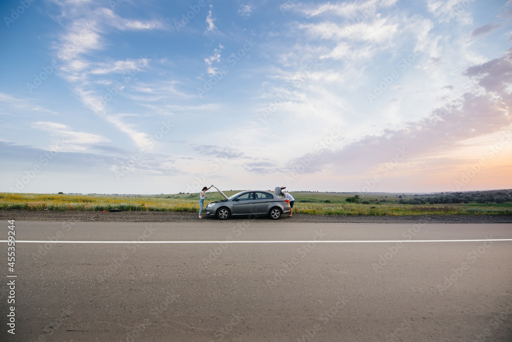A young girl stands near a broken car in the middle of the highway during sunset and tries to repair it. Breakdown and repair of the car. Troubleshooting the problem. A young girl stands near a broken car in the middle of the highway during sunset and tries to repair it. Breakdown and repair of the car. Troubleshooting the problem.