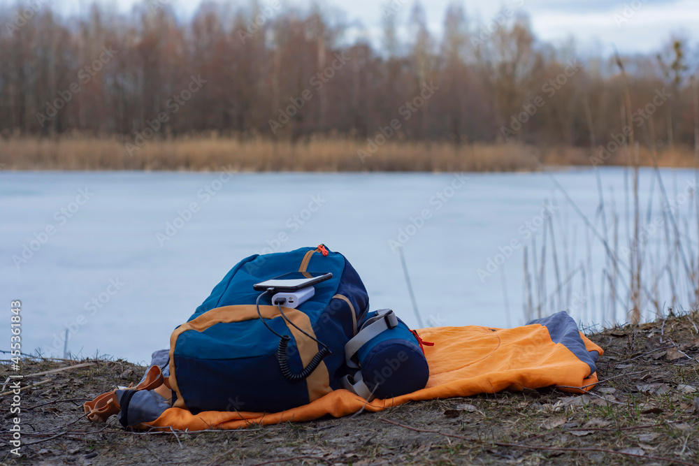 Smartphone is charged using a portable charger. Power Bank charges the phone outdoors with a backpack for tourism in the background of nature and the river. Smartphone is charged using a portable charger. Power Bank charges the phone outdoors with a backpack for tourism in the background of nature and the river.