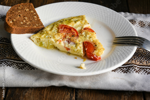 A slice of rustic omelet with potatoes and tomatoes on a white plate. A slice of bread, a kitchen napkin, and a wooden table.