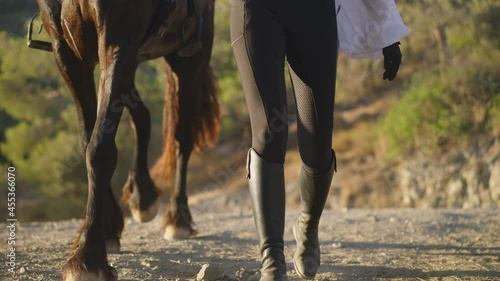 Front view legs of unrecognizable horse and woman strolling in slow motion on dusty mountain road. Slim Caucasian equestrian and purebred graceful animal walking in sunlight. Live camera photo