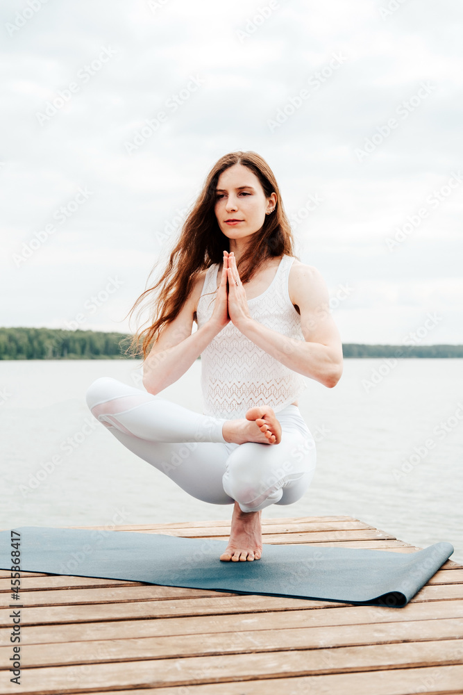 Beautiful young woman in white sports clothes is sitting on  wooden pier on lake in  balance yoga position with namaste gesture.