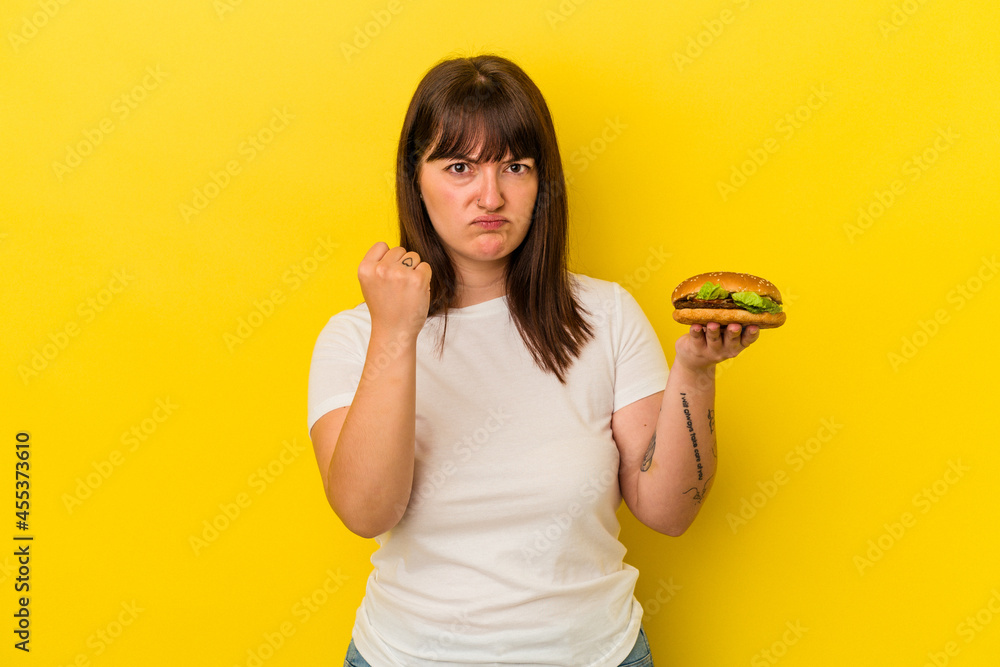 Young curvy caucasian woman holding burger isolated on yellow background showing fist to camera, aggressive facial expression. Young curvy caucasian woman holding burger isolated on yellow background showing fist to camera, aggressive facial expression.