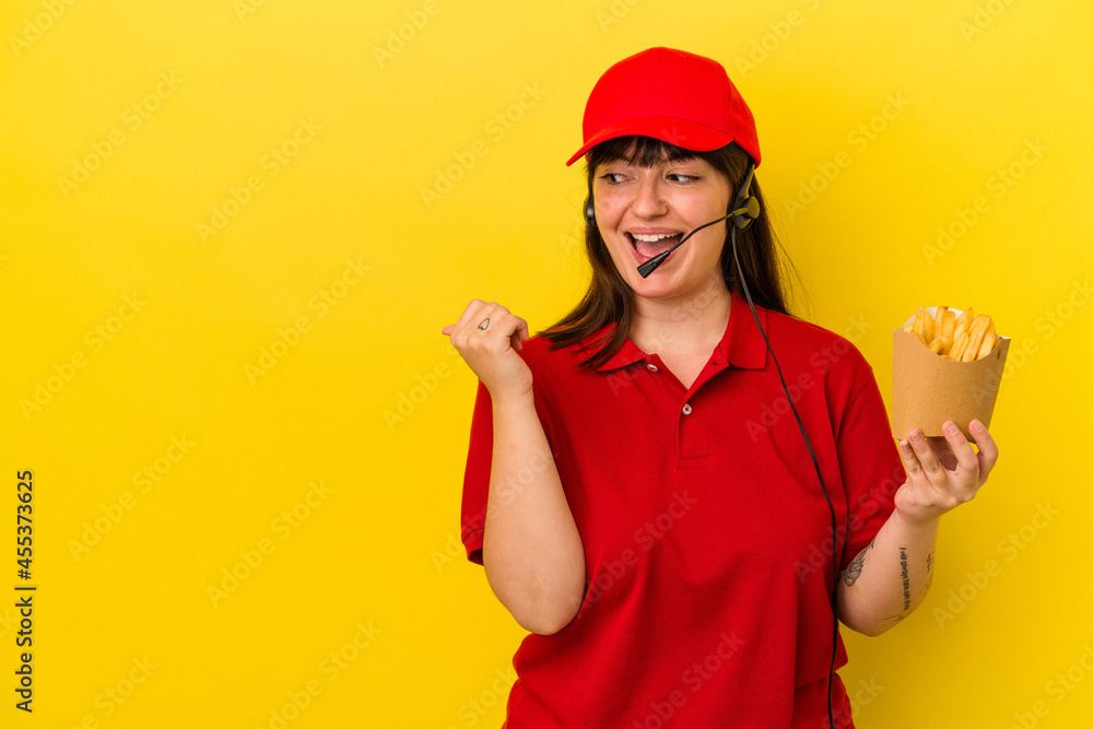 Young curvy caucasian woman fast food restaurant worker holding fries isolated on blue background points with thumb finger away, laughing and carefree.