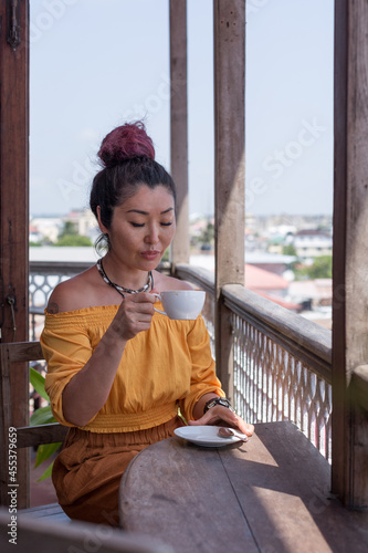Beautiful brunette girl of Asian appearance in an orange blouse drinks coffee on the balcony