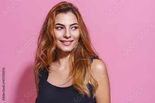 Seductive charming woman in black dress. On pink background. Smiling. With long brown hair looking away 