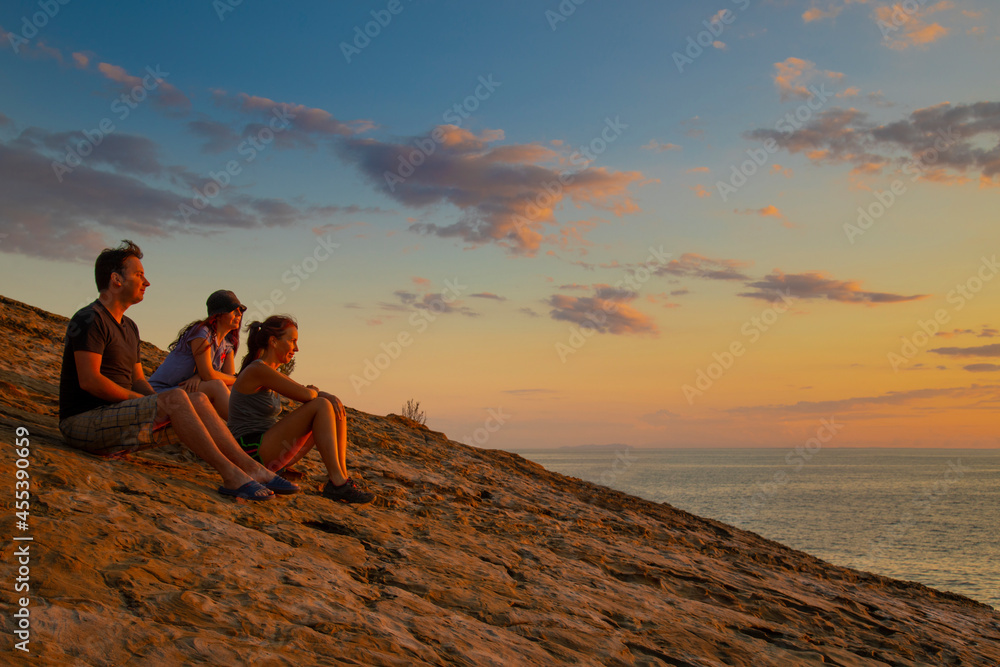 friends two women and a man at sunset time sit on a mountain above the sea and look into the distance