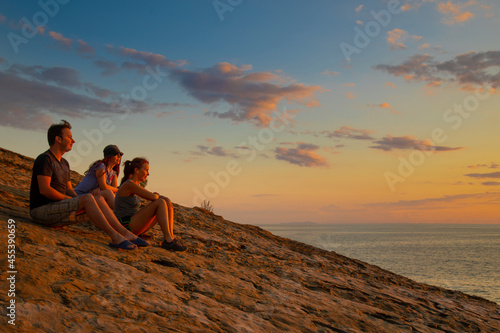 friends two women and a man at sunset time sit on a mountain above the sea and look into the distance