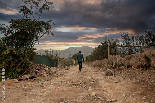 A man walks along a dirt and stone road, with a spectacular sky in the background.