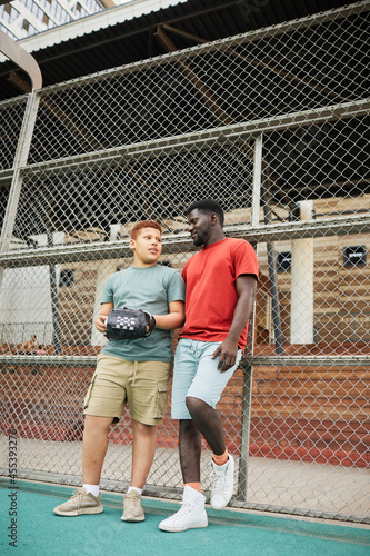 Teenage African American baseball player with glove standing against metal fence and chatting with father