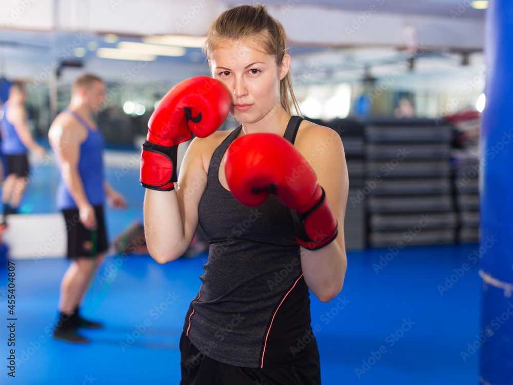 Portrait of young positive female boxer in red boxing gloves training in fitness center