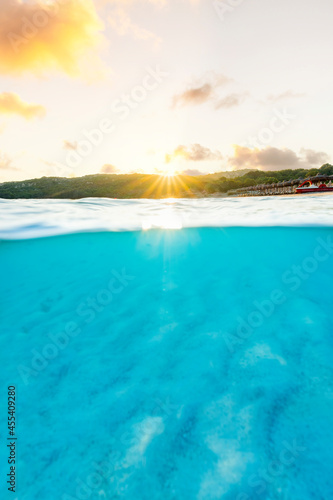 (Selective focus) Split shot, over under picture. Crystal clear, turquoise water and some beach umbrellas illuminated during a stunning sunrise. Grande Pevero Beach, Sardinia, Italy. © Travel Wild