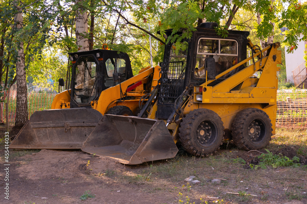 Two mini loaders with a bucket stand in the shade of a tree Two mini loaders with a bucket stand in the shade of a tree