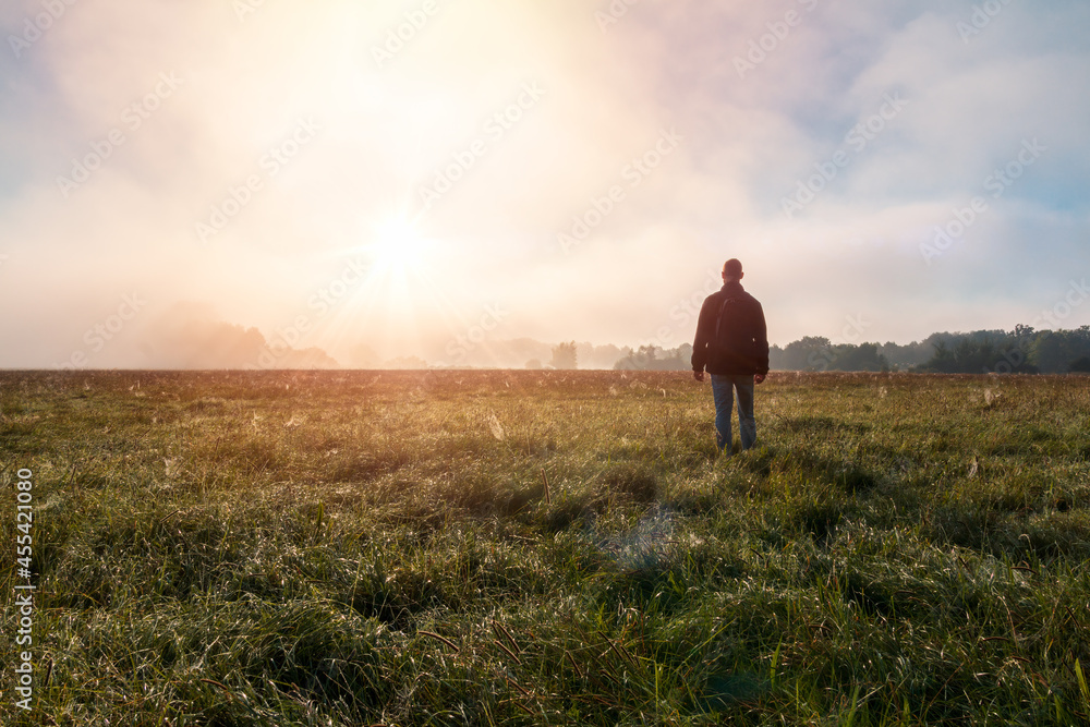 Young caucasian man standing, walking on meadow in misty fog at sunrise. Czech summer landscape