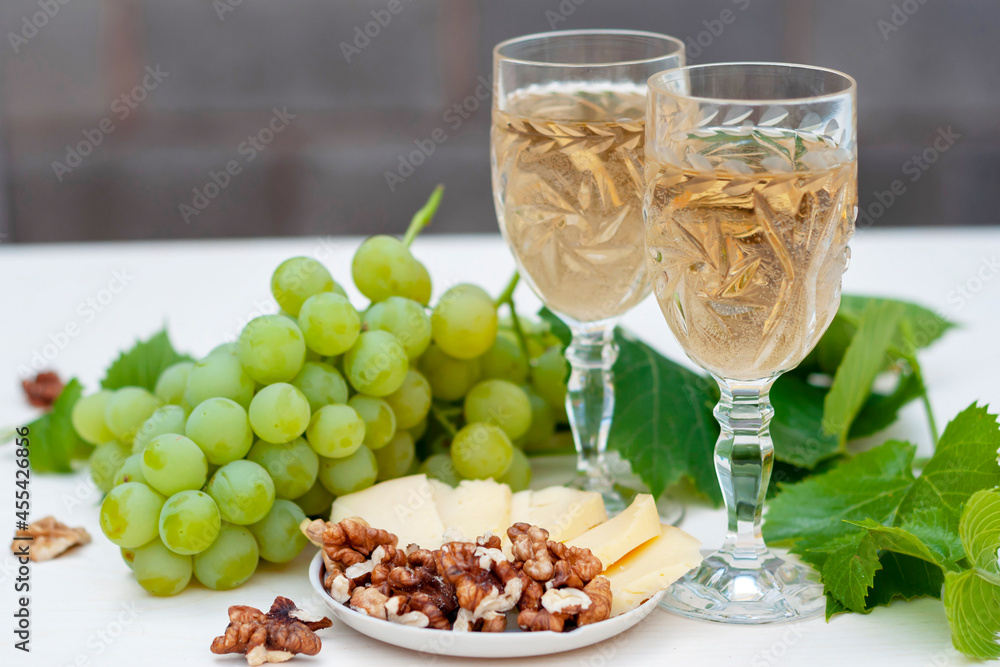 Two glasses of white wine, bunches of grapes, a young vine, cheese and walnuts on a light wooden background.