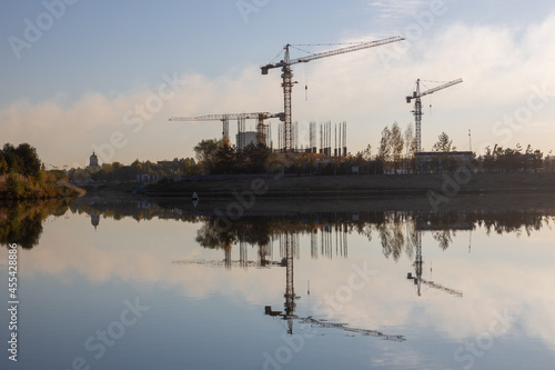 Construction site with cranes and buildings with their reflection in the water. Ishim river, Nur-Sultan, Kazakhstan.