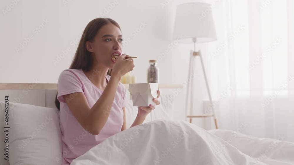 Food delivery service. Young positive woman eating chinese noodles from box with chopsticks, sitting in bed in morning Food delivery service. Young positive woman eating chinese noodles from box with chopsticks, sitting in bed in morning