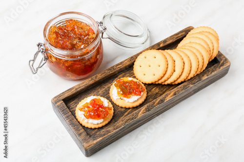 Delicious jalapeno pepper jam in jar and wooden board with crackers on white background photo