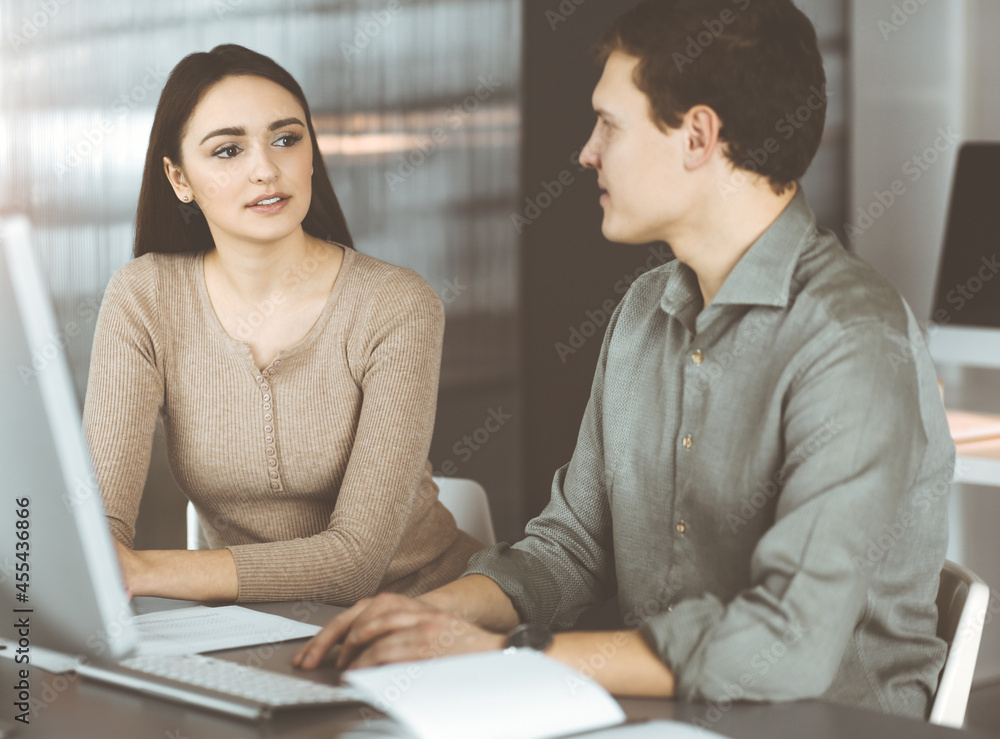 Young business people are talking to each other, while sitting at the desk in sunny office. Businesswoman is explaining something to her colleague Young business people are talking to each other, while sitting at the desk in sunny office. Businesswoman is explaining something to her colleague