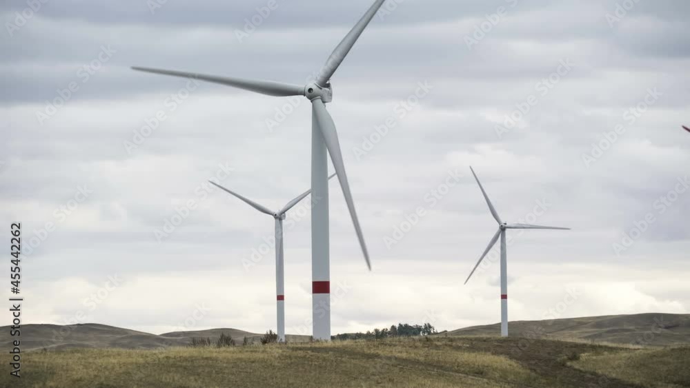 Motion the blades of a large wind turbine in a field against a background of cloudy grey sky on the horizon with a beautiful hills. Alternative energy sources. Windy park. Ecological energy.Industrial Motion the blades of a large wind turbine in a field against a background of cloudy grey sky on the horizon with a beautiful hills. Alternative energy sources. Windy park. Ecological energy.Industrial