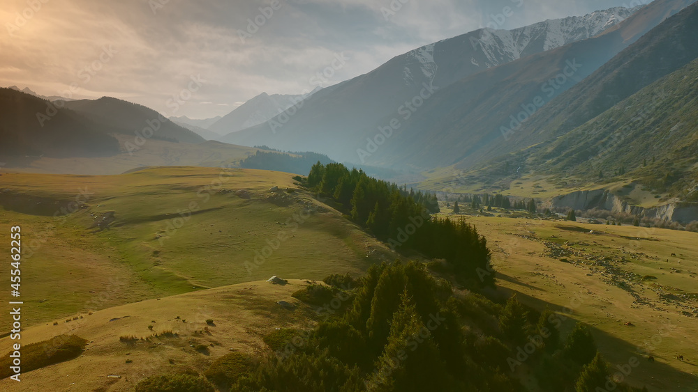 Aerial flight over green hills and fields in the rays of sunset sunlight, against the background of huge mountains with glaciers. A picturesque idyllic landscape with a light fog, a river, a hill.