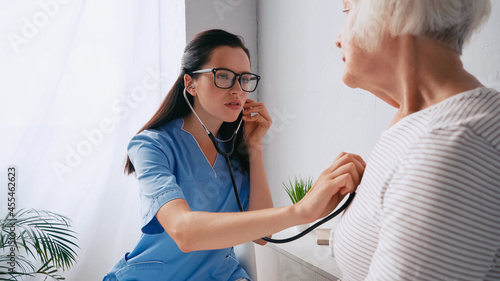brunette nurse in eyeglasses examining senior woman with stethoscope