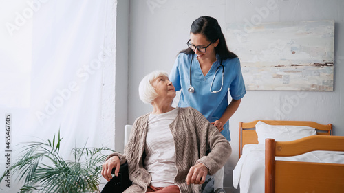 happy nurse in eyeglasses moving aged woman in wheelchair photo