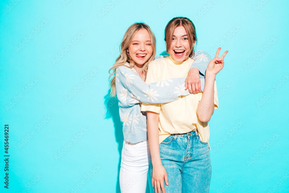 Two young beautiful blond smiling hipster female in trendy summer clothes. Sexy carefree women posing near blue wall in studio. Trendy and positive models having fun.Shows peace sign Two young beautiful blond smiling hipster female in trendy summer clothes. Sexy carefree women posing near blue wall in studio. Trendy and positive models having fun.Shows peace sign
