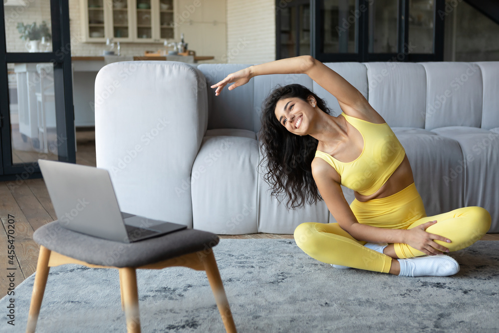 Fototapeta premium Fit young woman doing yoga in front of laptop, bending aside during home workout, copy space