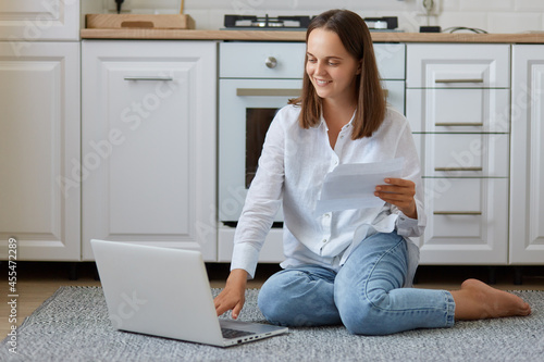 Free Download Happy young Caucasian woman sitting on the floor in kitchen, holding paper document or letter, checking domestic bills, smiling, doing paperwork work or study at home. - GFXLoad