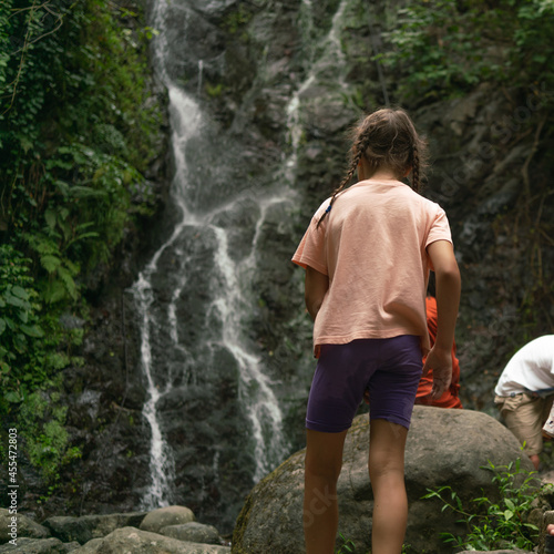 A little girl looks at the streams of a waterfall rapidly flowing along the steep surface of the mountain on a summer evening