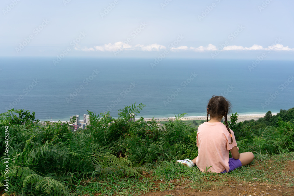 A little girl on a summer sunny day sits on a mountain above the sea and the beach and looks into the distance A little girl on a summer sunny day sits on a mountain above the sea and the beach and looks into the distance